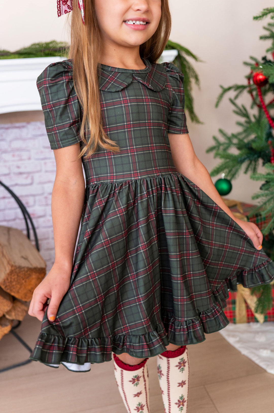 A young girl wearing a dark green plaid dress with red and white stripes, featuring a Peter Pan collar and short puff sleeves. She is standing in a cozy indoor setting with a Christmas tree and fireplace in the background.