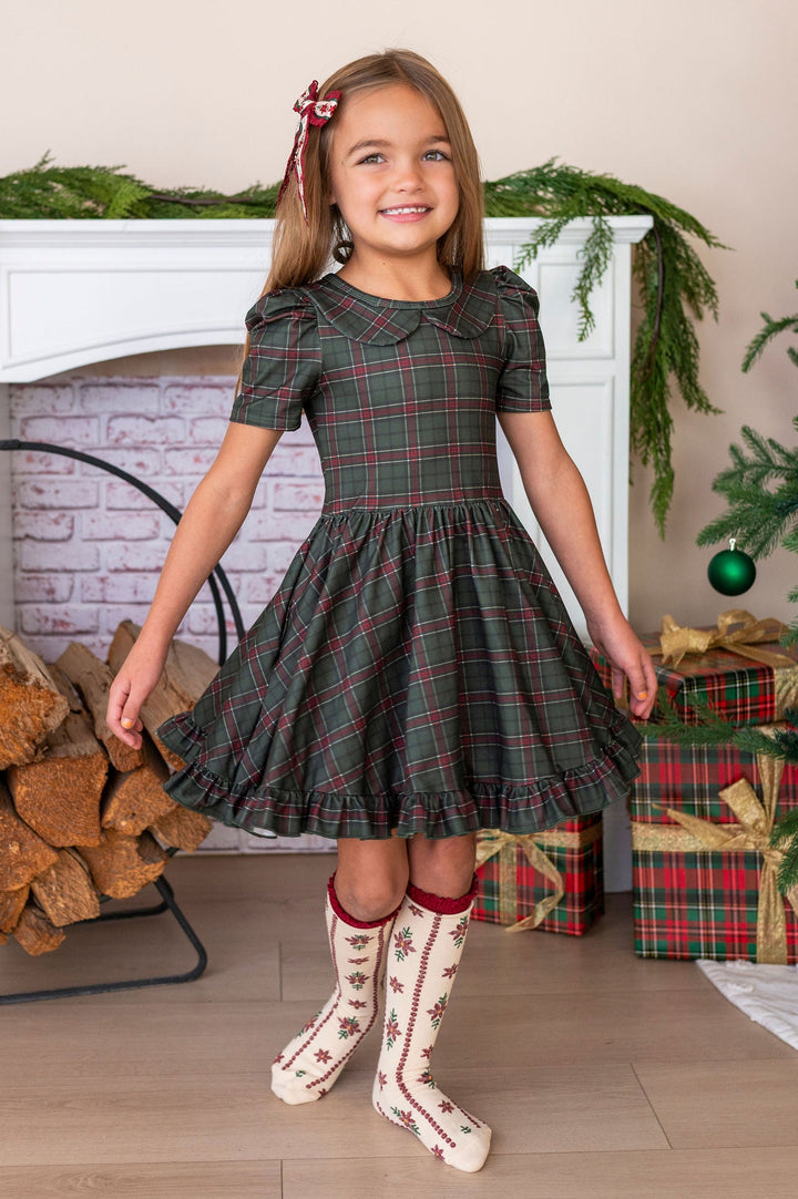 A young girl stands in a holiday-themed room wearing a short-sleeve plaid dress with a Peter Pan collar, cream socks with red trim, and a red hair bow. The background features a white fireplace mantel with green garland, a stack of firewood, wrapped Christmas presents, and a decorated Christmas tree.