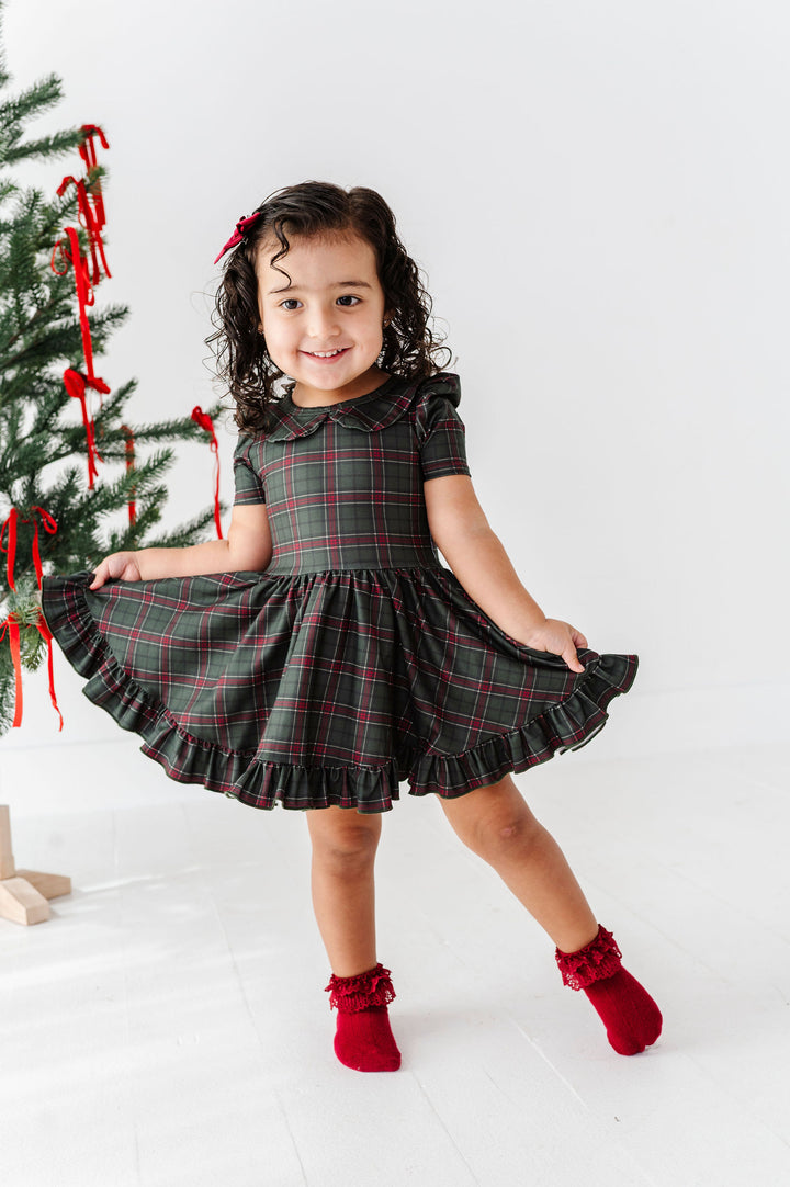 A young girl with curly dark hair wearing a dark green and red plaid short-sleeve dress with a ruffled collar and hem, paired with red ruffled socks. She is posing in a bright studio setting next to a decorated Christmas tree.