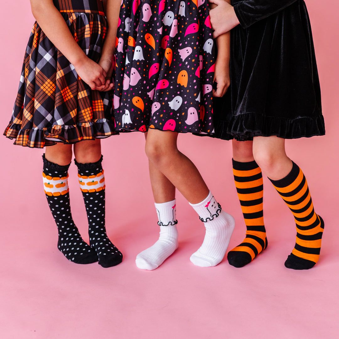 three girls holding hands in pumpkin dot knee socks, ghost midi socks and black and orange striped knee highs paired with coordinating Halloween dresses