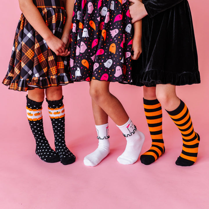 three girls holding hands in pumpkin dot knee socks, ghost midi socks and black and orange striped knee highs paired with coordinating Halloween dresses