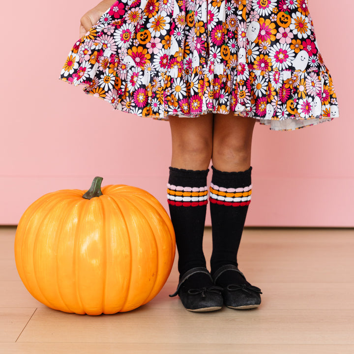 little girl standing by pumpkin in velvet floral Halloween print dress and black scallop trimmed knee high socks with stripes in matching colors