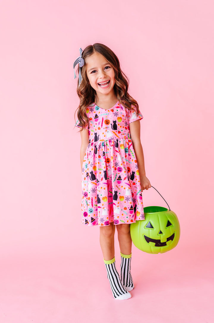 little girl holding green pumpkin bucket in pink Halloween themed twirl dress paired with black and white striped midi socks and hair bow