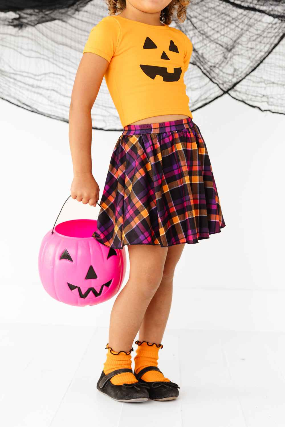 little girl holding pink pumpkin bucket in orange, purple, pink and black Halloween plaid skort, orange top with jack-o-lantern design and matching orange midi socks