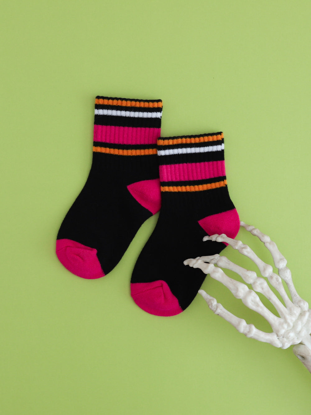 flat lay of black crew socks with pink and orange stripes on green background with skeleton hand