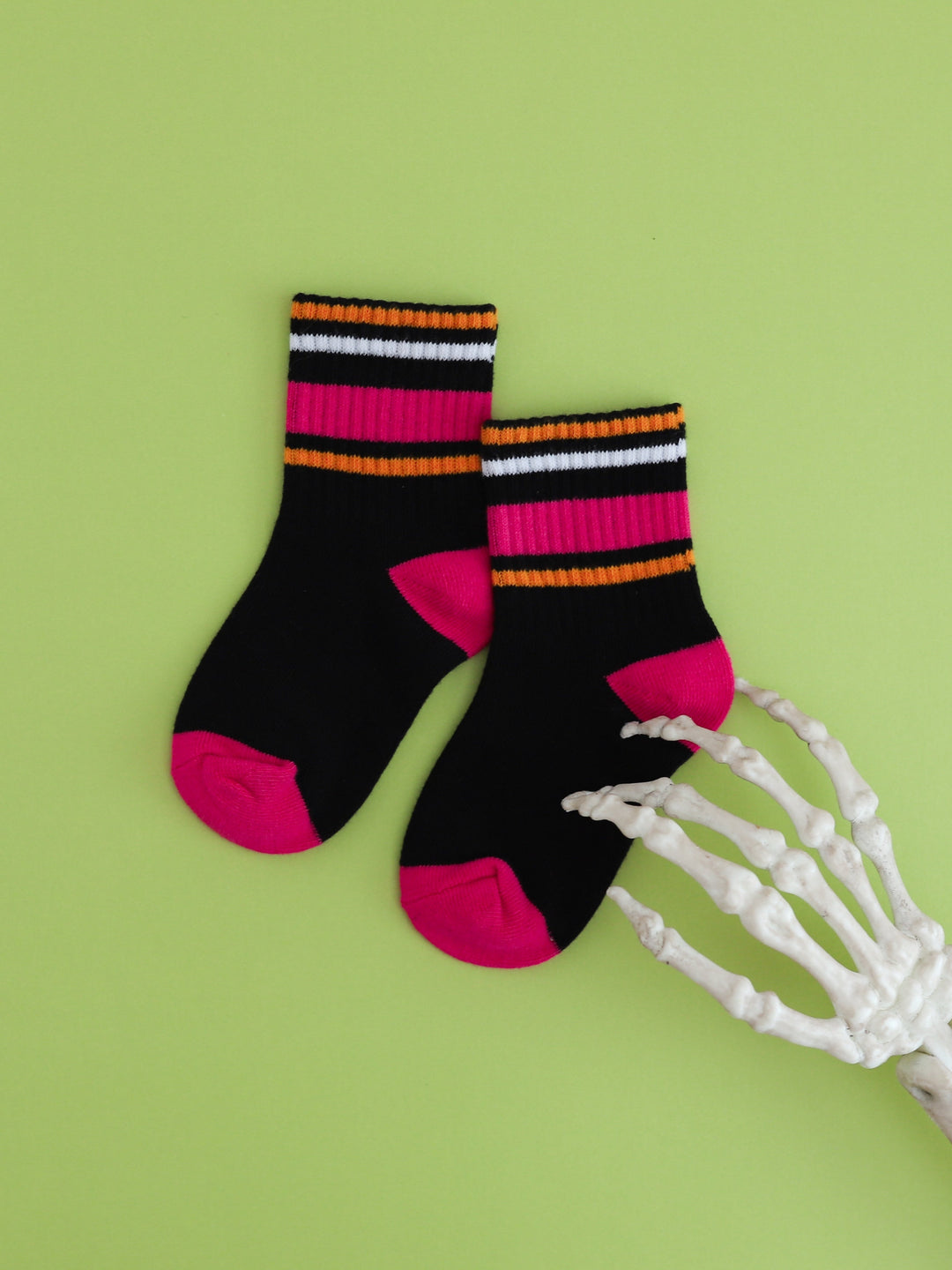 flat lay of black crew socks with pink and orange stripes on green background with skeleton hand