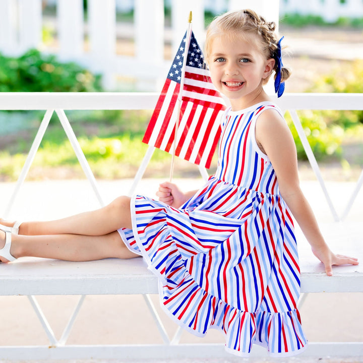 little girl sitting on white bench wearing 4th of july striped dress and holding american flag in her hand