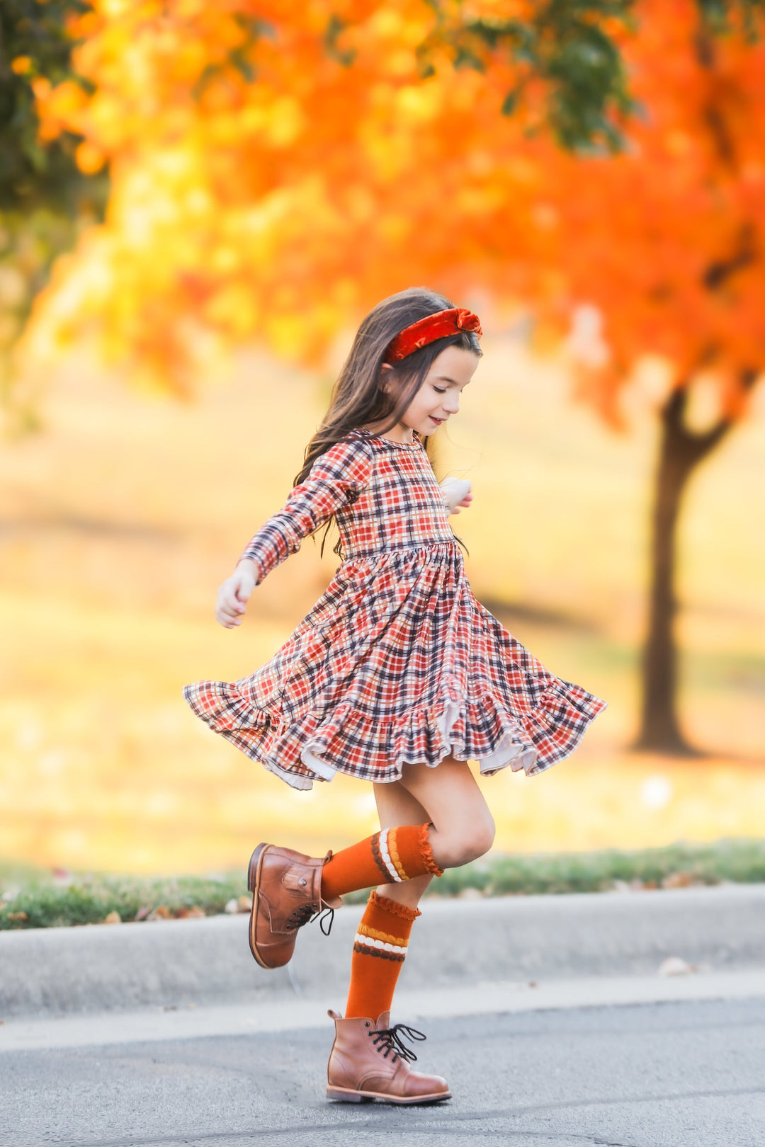 little girl in front of fall foliage twirling in fall plaid long sleeve velvet dress and matching pumpkin spice knee highs and headband