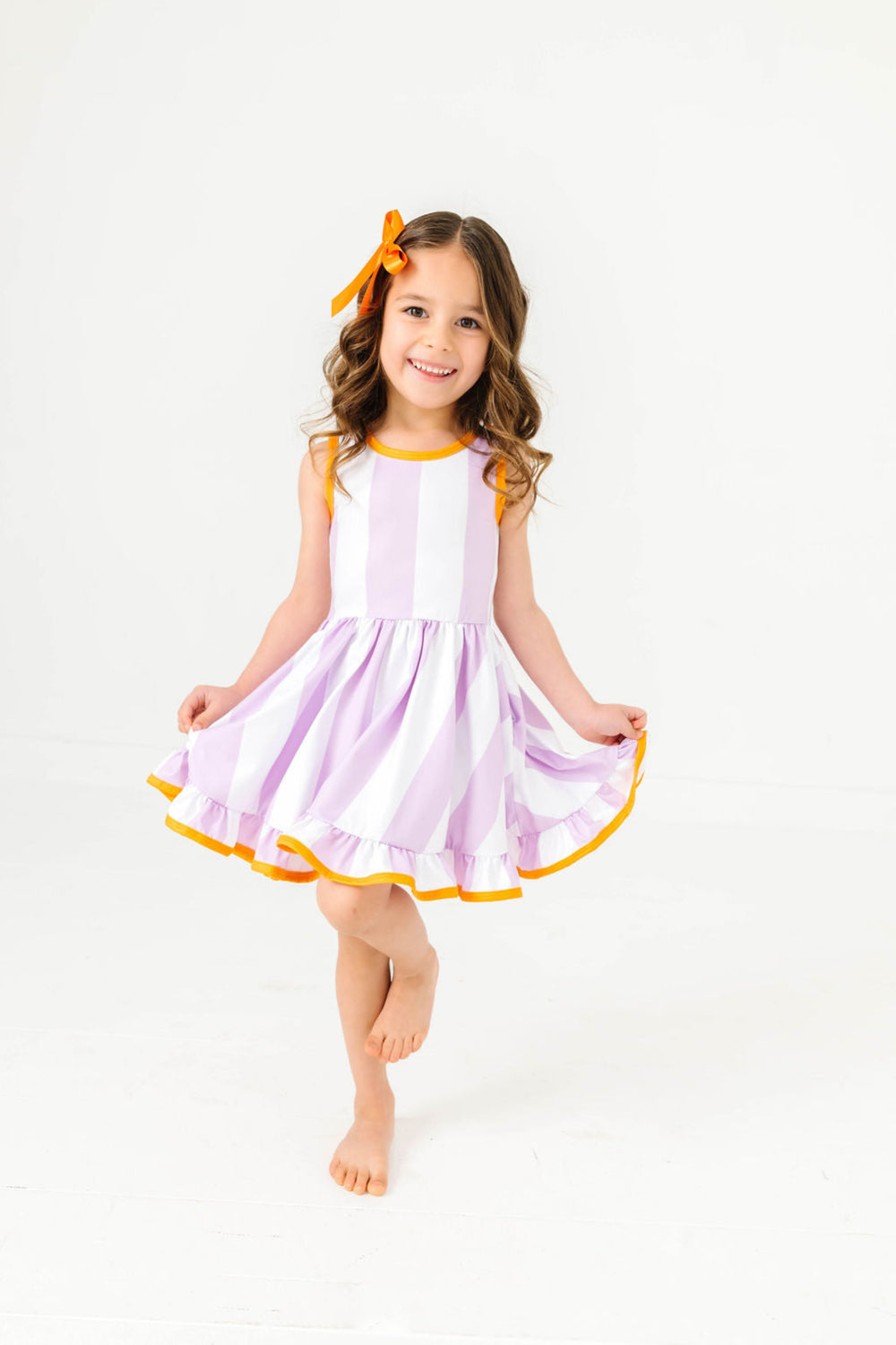 little girl in purple and white wide striped sleeveless twirl dress with orange trim and matching orange satin hair bow