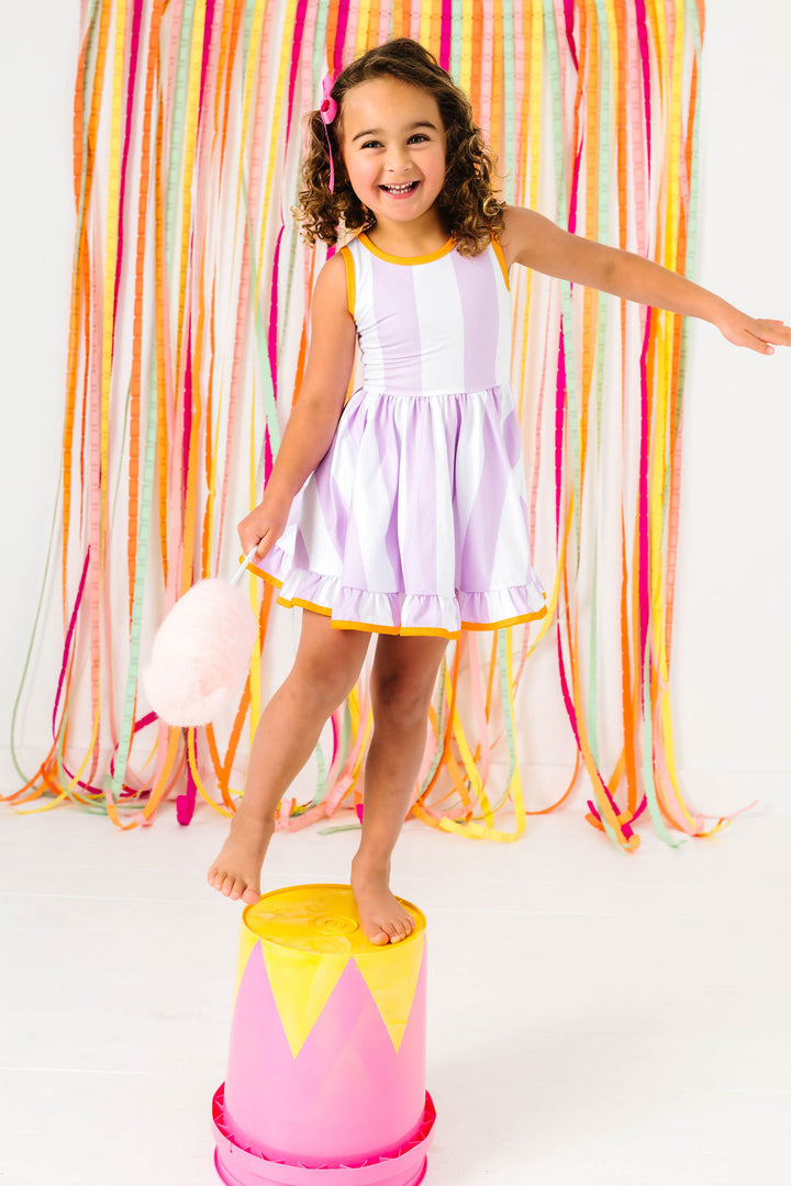 little girl standing on circus platform with cotton candy wearing light purple and white striped sleeveless twirl dress with orange trim