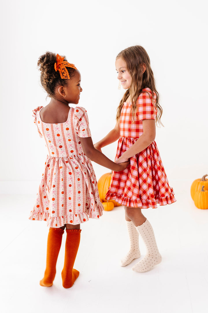 two girls holding hands and twirling in pumpkin wallpaper and pumpkin plaid square neck twirl dresses paired with matching knee socks and bows in orange and cream