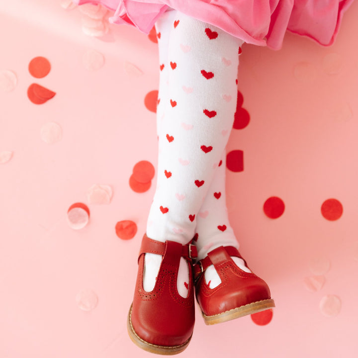 little girl sitting in white knit tights with pink and red tiny heart pattern, pink twirl dress and red shoes