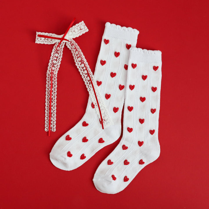 flat lay photo of girls white scallop knee high socks with red heart design and matching white lace, red ribbon bow