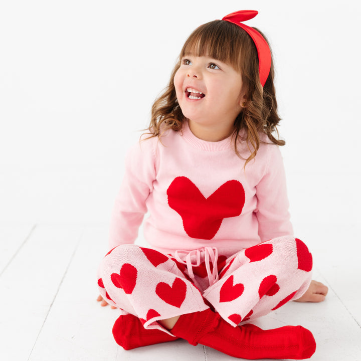 little girl sitting in pink with red hearts cotton knit sweater pant set and matching red socks and satin headband