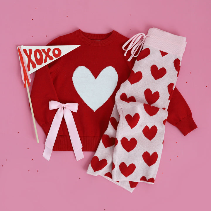 styled Valentine's flat lay of red knit sweater with white heart, pink sweater pants with red hearts and matching light pink satin bow