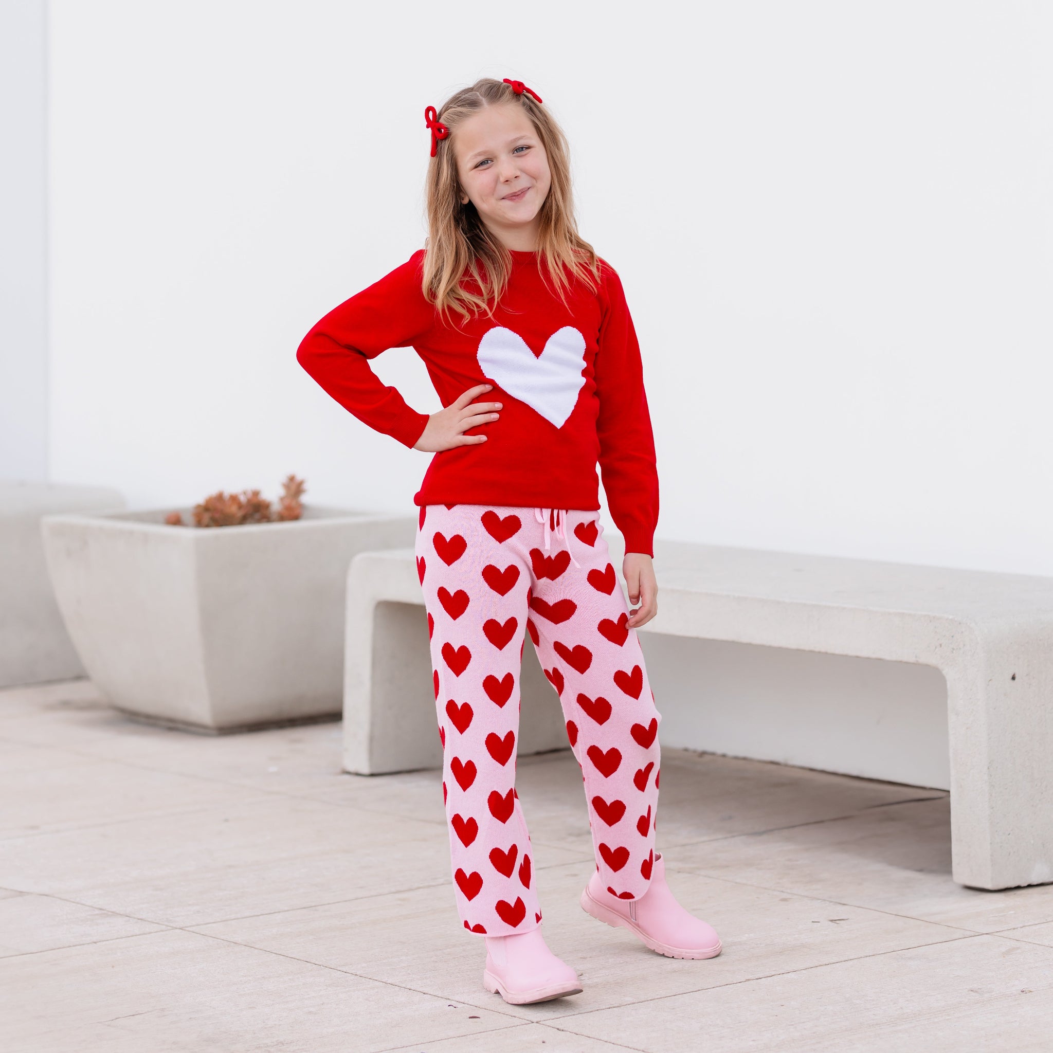 girl in Valentines Day cotton knit sweater set featuring red pullover with white heart, pink pants with red hearts and matching red yarn pigtail bows