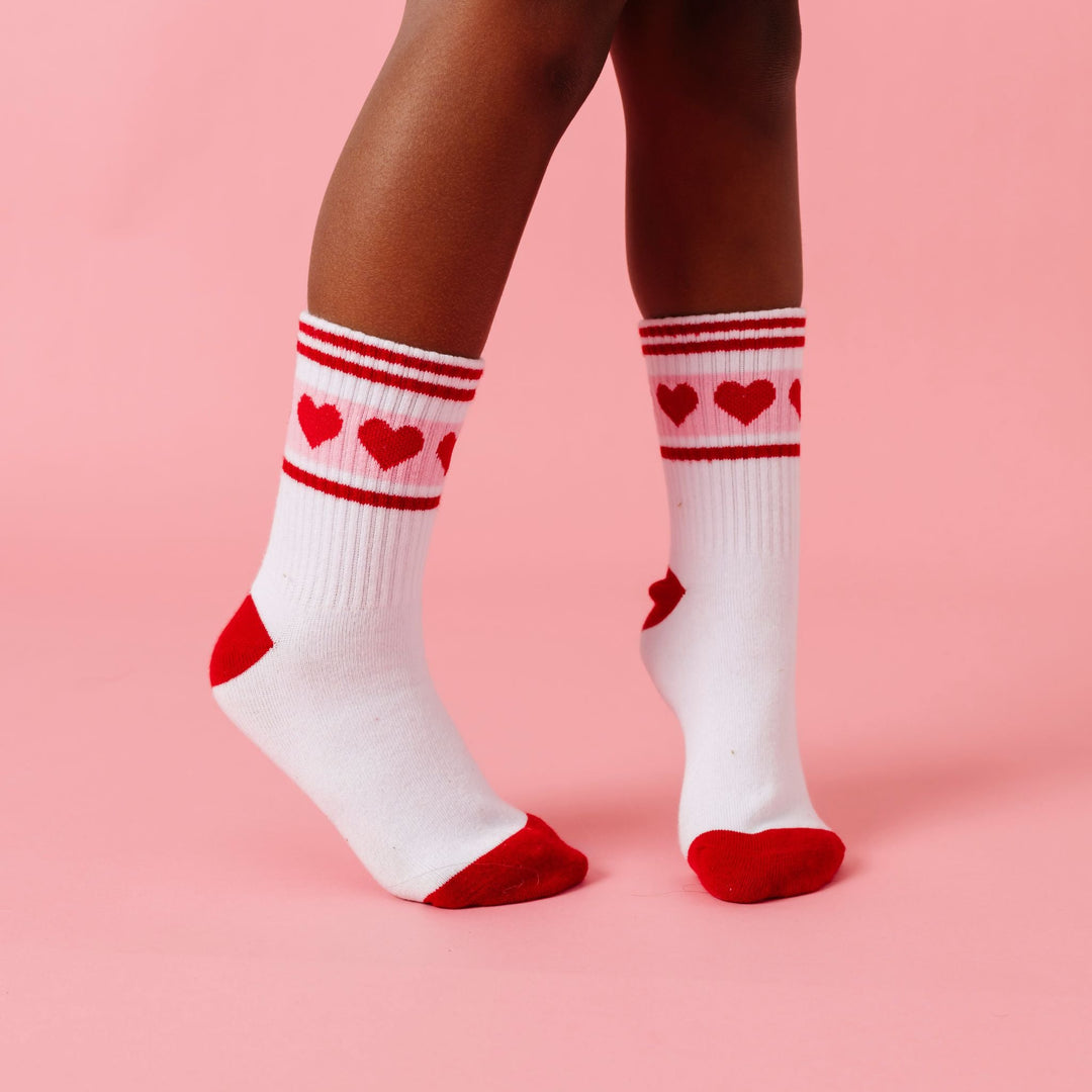 closeup of child wearing white crew socks with red toe and heel detail, red stripes and red hearts on pink 