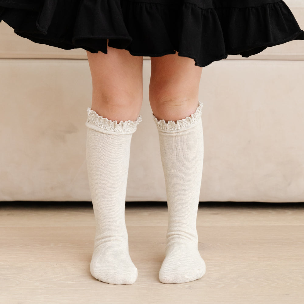 little girl standing in black dress wearing light heathered ivory melange lace top knee high socks