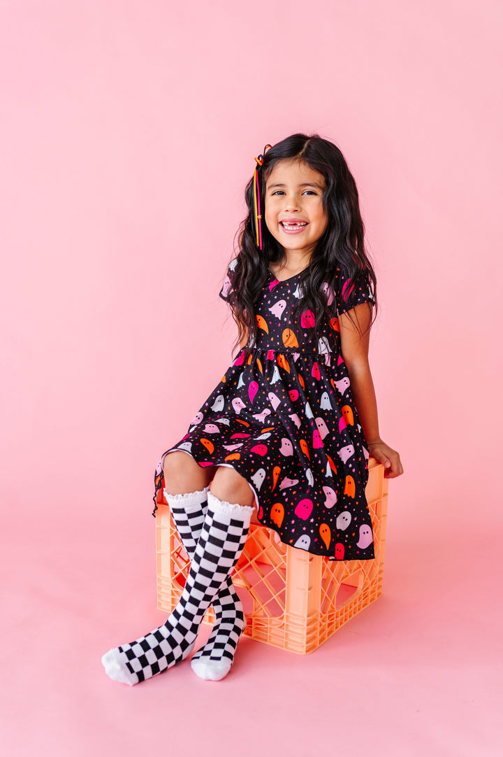 young girl sitting on orange crate wearing black with colorful ghost print twirl dress paired with black and white checkered lace top knee socks