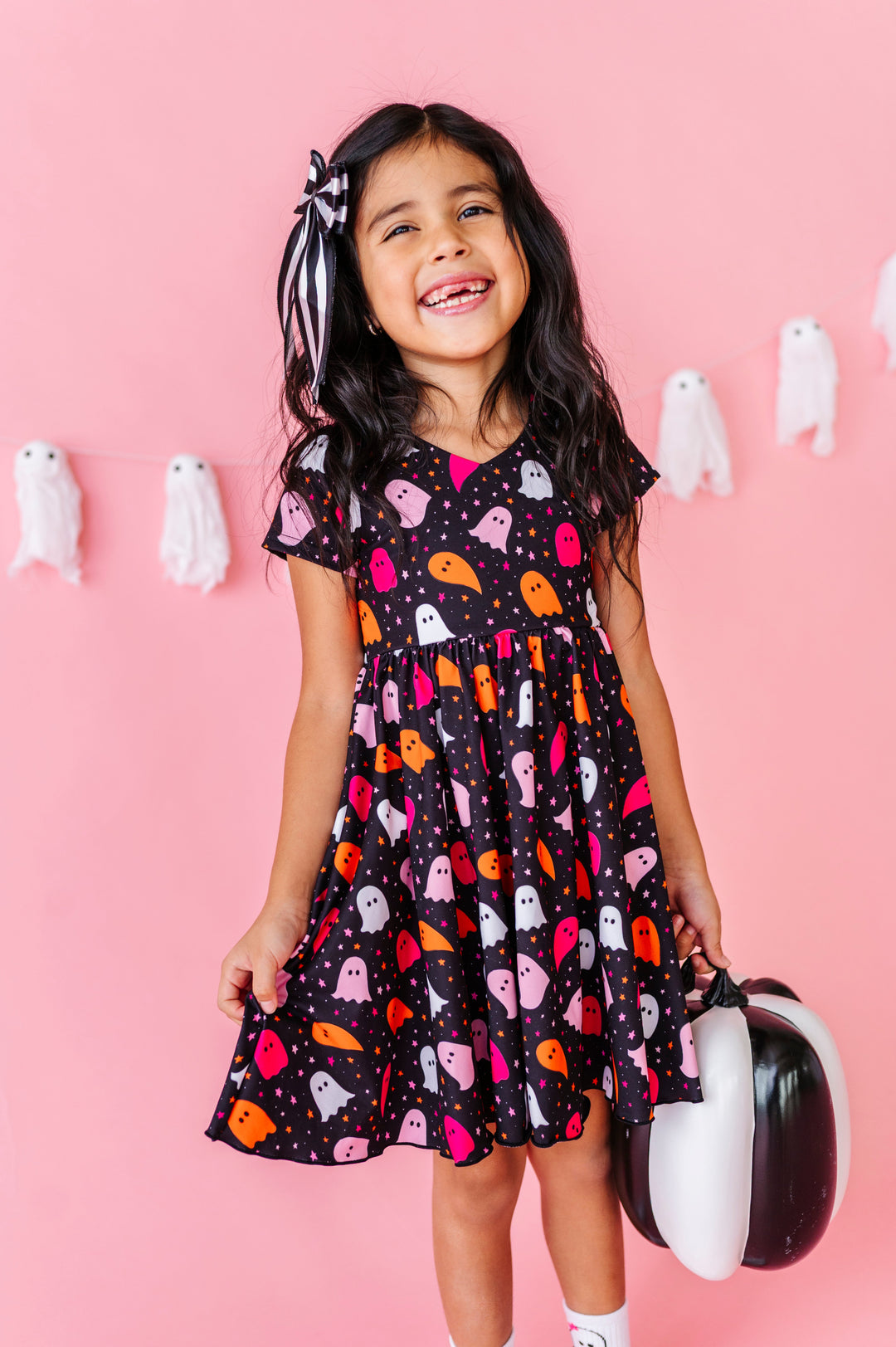 young girl in front of ghost decor holding black and white pumpkin in black v-neck twirl dress with colorful ghost print