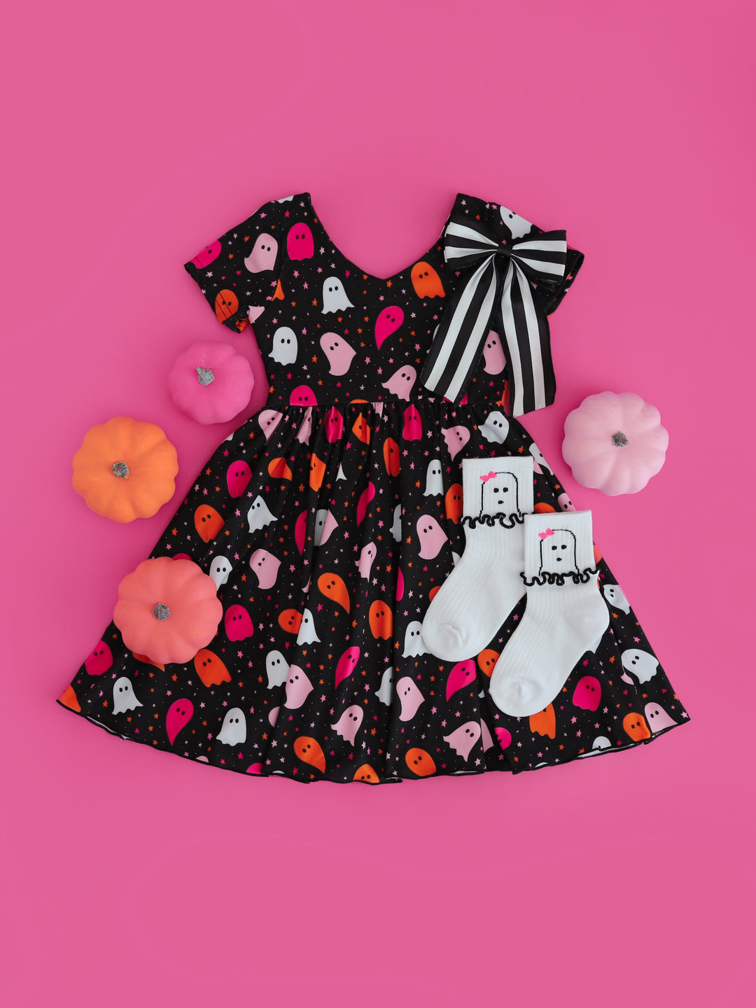 girls styled black with ghost print v-neck twirl dress paired with black and white striped hair bow and ghost midi socks on pink background with pink mini pumpkins
