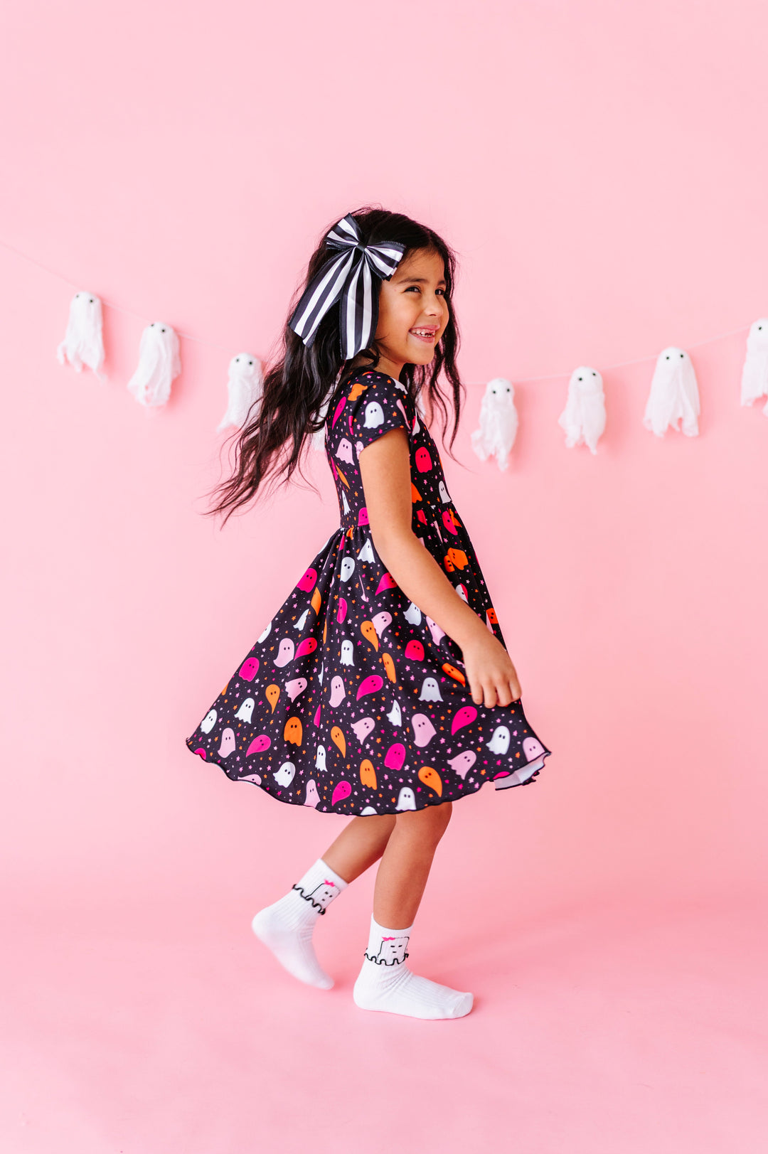 young girl twirling in black short sleeve dress with pink, white and orange ghost and star print paired with black and white striped hair bow and white midi socks with ruffled ghost design at ankle