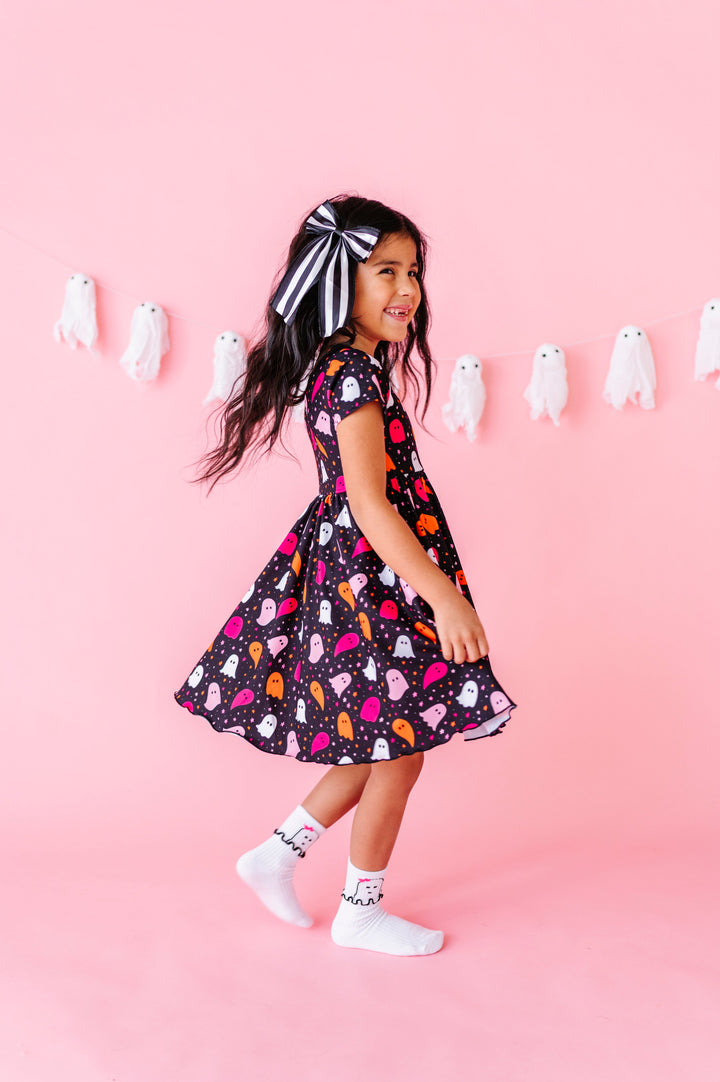 young girl twirling in black short sleeve dress with pink, white and orange ghost and star print paired with black and white striped hair bow and white midi socks with ruffled ghost design at ankle