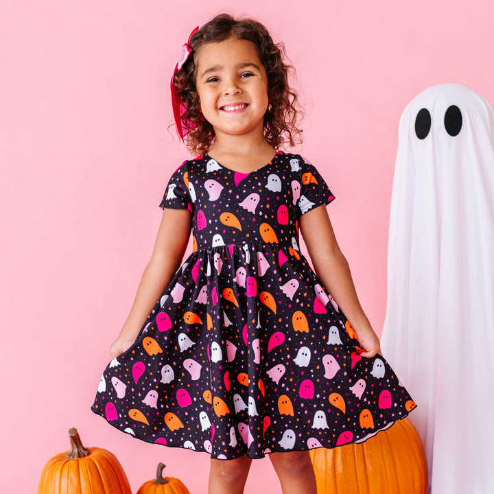 little girl standing by pumpkins and sheet ghost wearing black v-neck twirl dress with orange and oink ghost and star design for Halloween