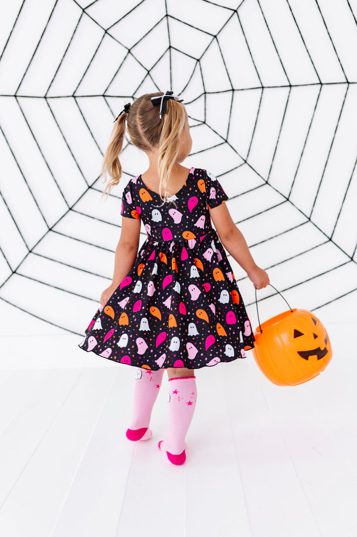 young girl twirling in front of giant spider web decoration with orange pumpkin bucket wearing black v-neck twirl dress with pink, orange and white ghost print paired with matching pink ghost knee socks