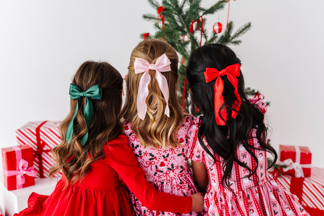 Three young girls viewed from behind, sitting together in front of a Christmas tree with wrapped presents. The girls wear red and pink holiday dresses and have large hair bows in green, pink, and red.