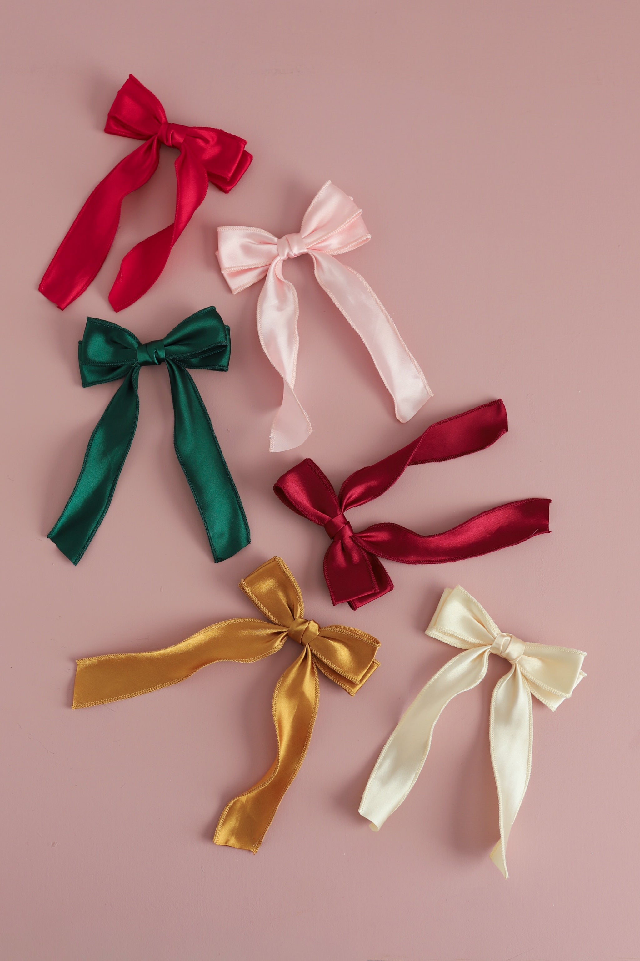 Six satin ribbon bows in assorted colors, including red, light pink, forest green, deep crimson, gold, and cream, arranged on a solid dusty pink background.