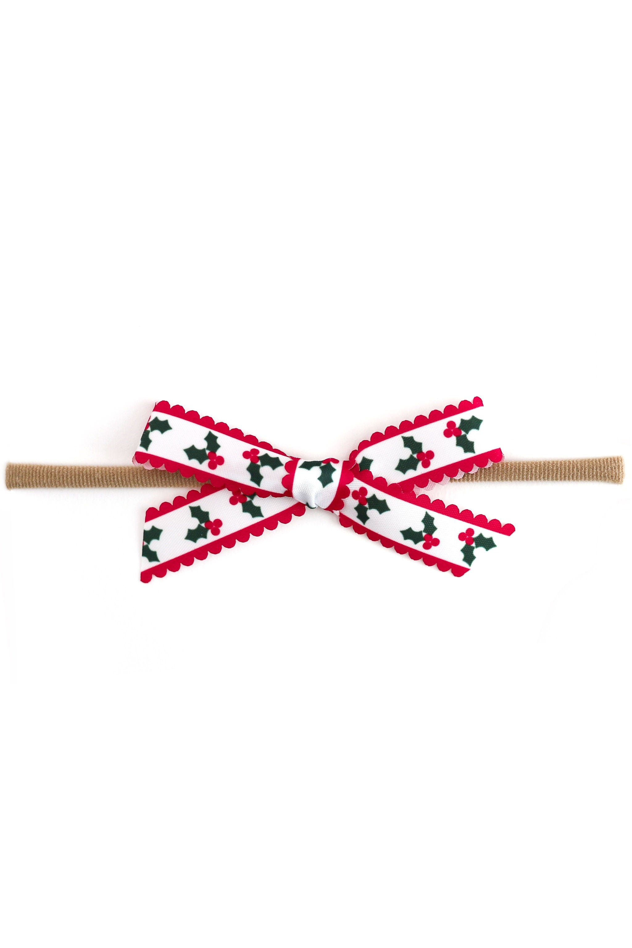 A festive baby bow headband featuring a white ribbon printed with a red and green holly pattern, finished with red scalloped trim, attached to a tan elastic band, shown on a plain white background.
