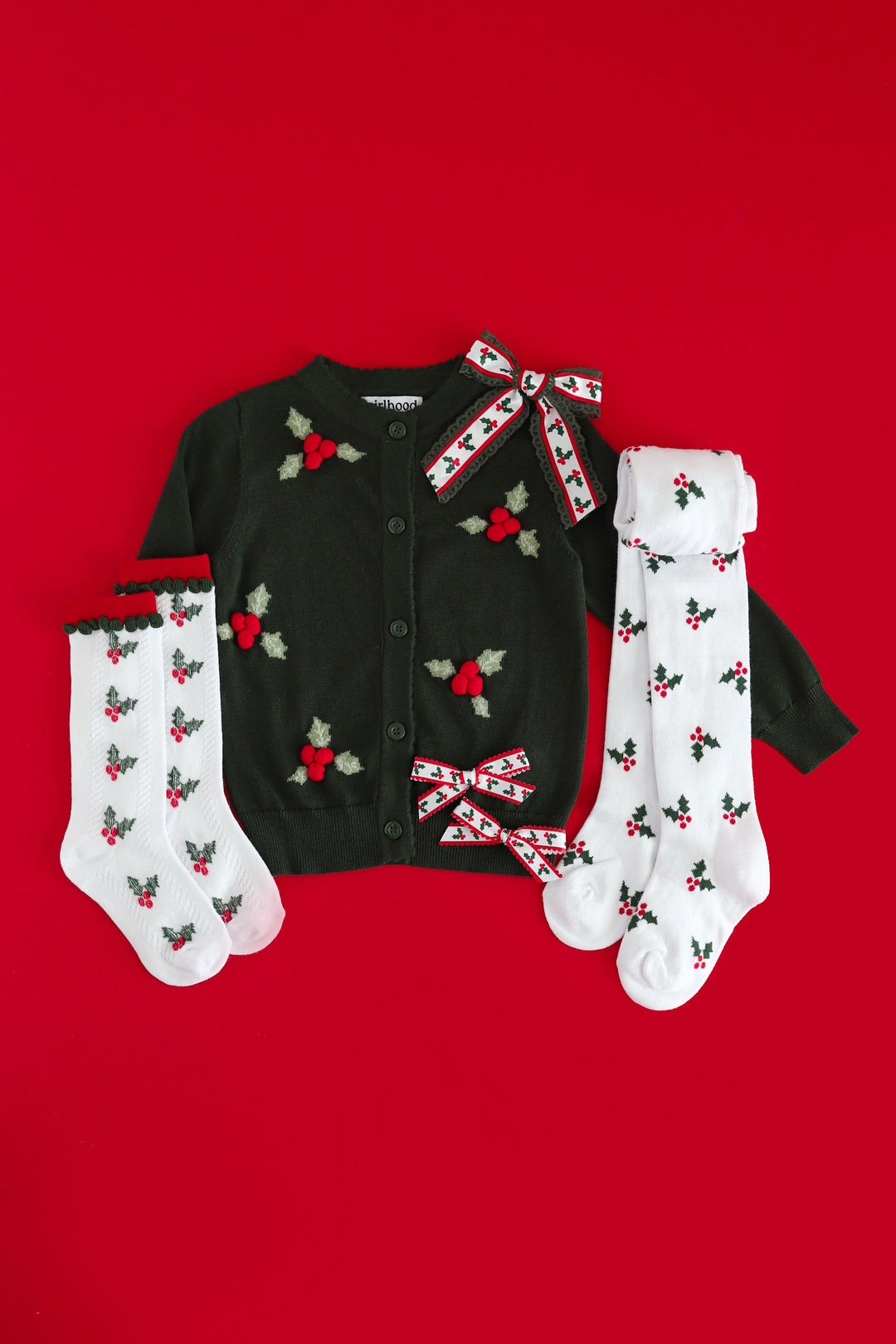 A Christmas-themed xmas outfit set featuring a dark green knit cardigan with red and green holly knit design, white knit socks with red and green holly prints, and matching red-and-white patterned bow ties, laid flat on a solid red background.