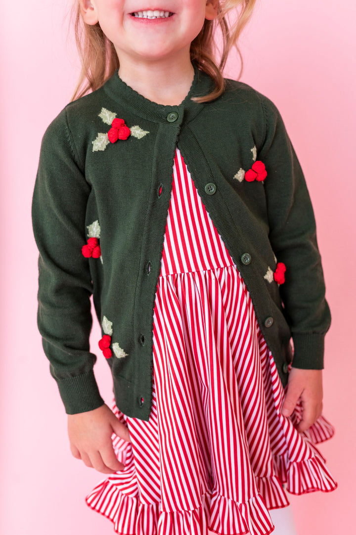 A young child wearing a dark green knit cardigan with red and green holly floral appliques over a red and white striped dress, set against a solid pink background.