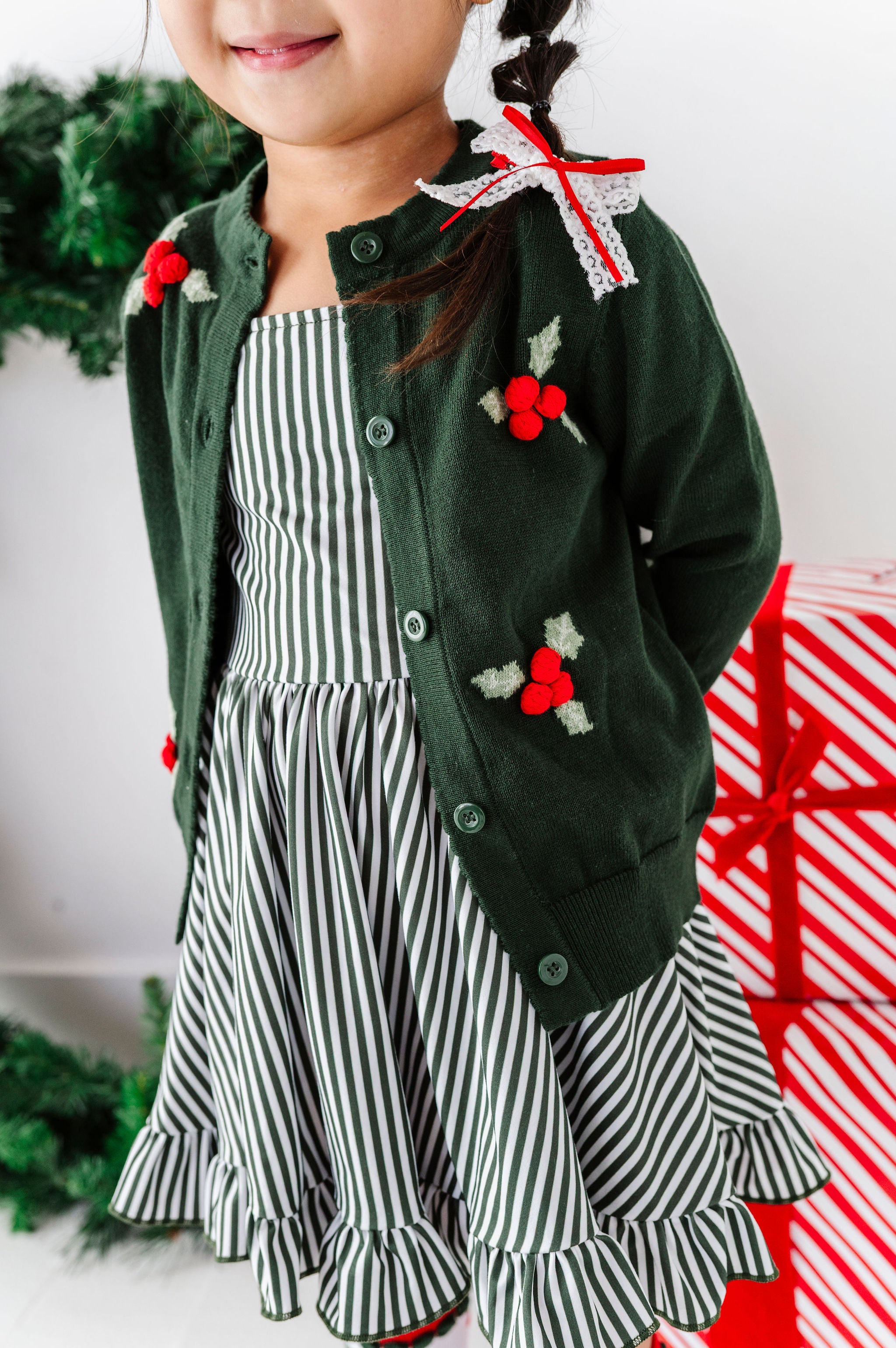A young girl wearing a festive holiday outfit featuring a dark green knit cardigan with red holly accents over a green and white striped dress, posed next to a wrapped Christmas gift.