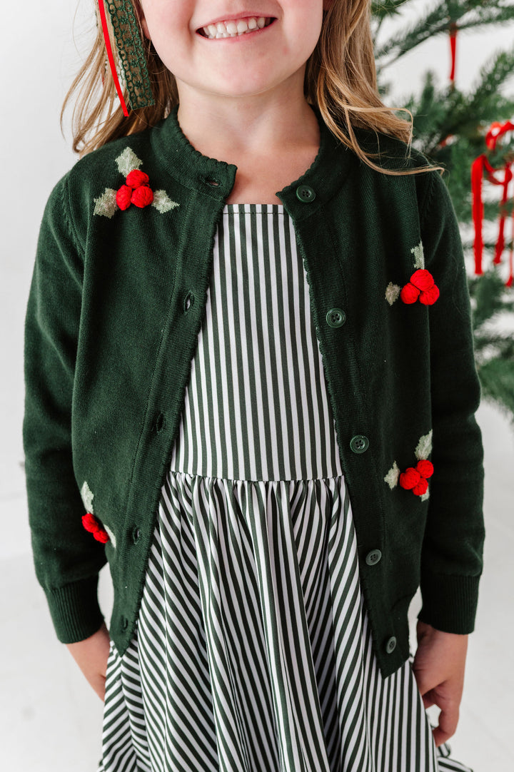 A young girl wearing a dark green knit cardigan with red berry and holly leaf knit design, layered over a green and white striped dress, with a Christmas tree visible in the background.