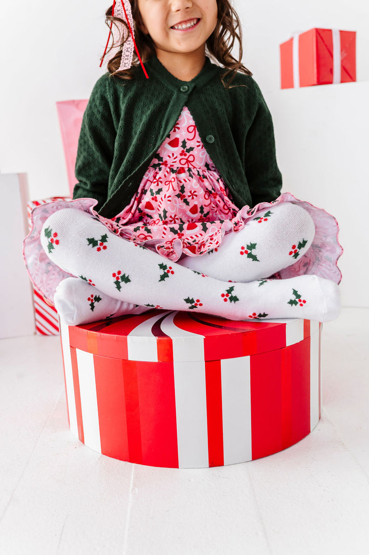 girl sitting on a christmas gift box wearing holly knit tights with matching christmas dress and sweater