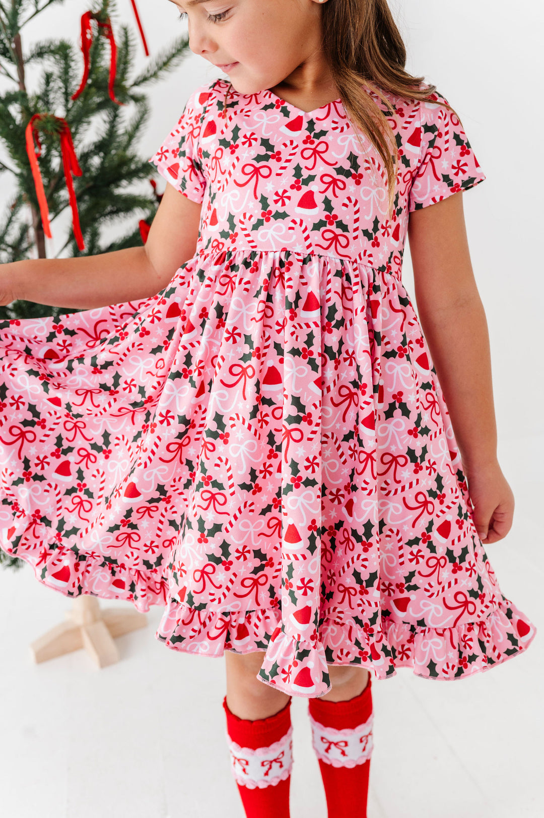 A young girl wearing a pink short-sleeve dress with a red and black Christmas candy cane, holly, and heart pattern, paired with red and white socks, standing in front of a decorated Christmas tree.
