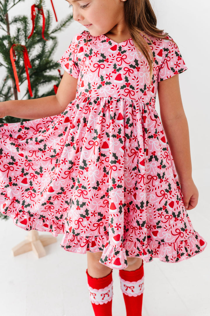 A young girl wearing a pink short-sleeve dress with a red and black Christmas candy cane, holly, and heart pattern, paired with red and white socks, standing in front of a decorated Christmas tree.