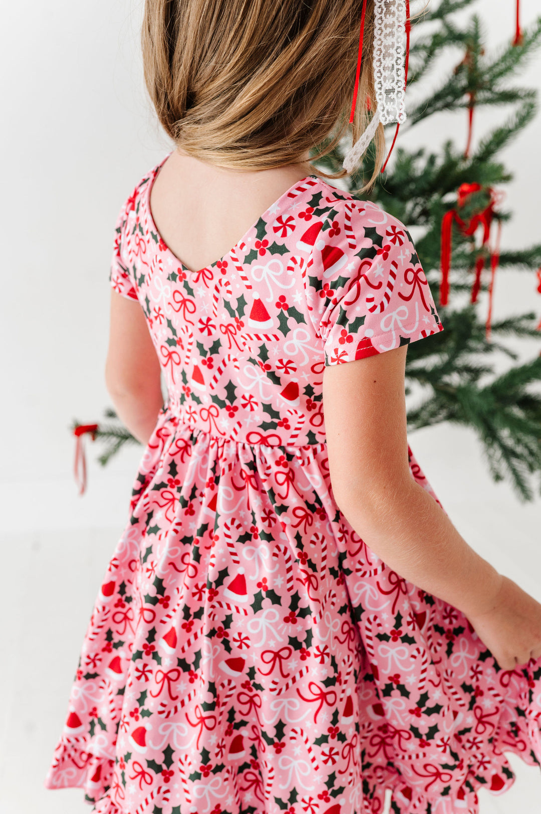 A young girl viewed from behind wearing a short-sleeve V-neck dress with a festive Christmas print of red bows, candy canes, and holly on a light pink background. She is standing next to a decorated Christmas tree.