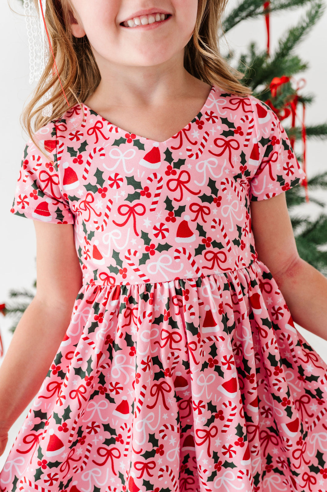A young girl wearing a pink short-sleeve dress with a festive Christmas print of red bows, candy canes, holly leaves, and bells, standing in front of a decorated Christmas tree.