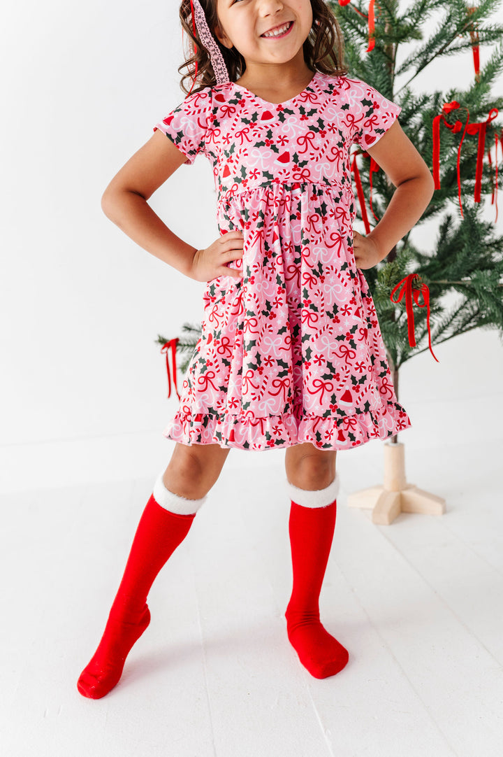 A young girl wearing a pink short-sleeve dress with a red and black holiday pattern, paired with bright red knee-high socks, standing in front of a decorated Christmas tree.