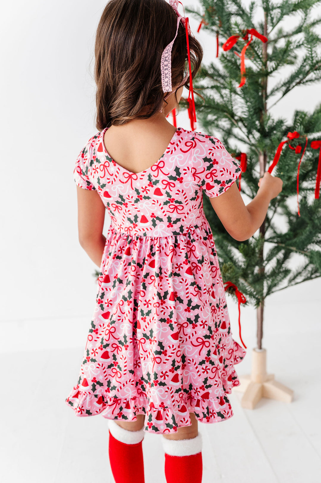 A young girl viewed from behind wearing a pink short-sleeve dress with a red and green holiday print, standing next to a decorated Christmas tree.