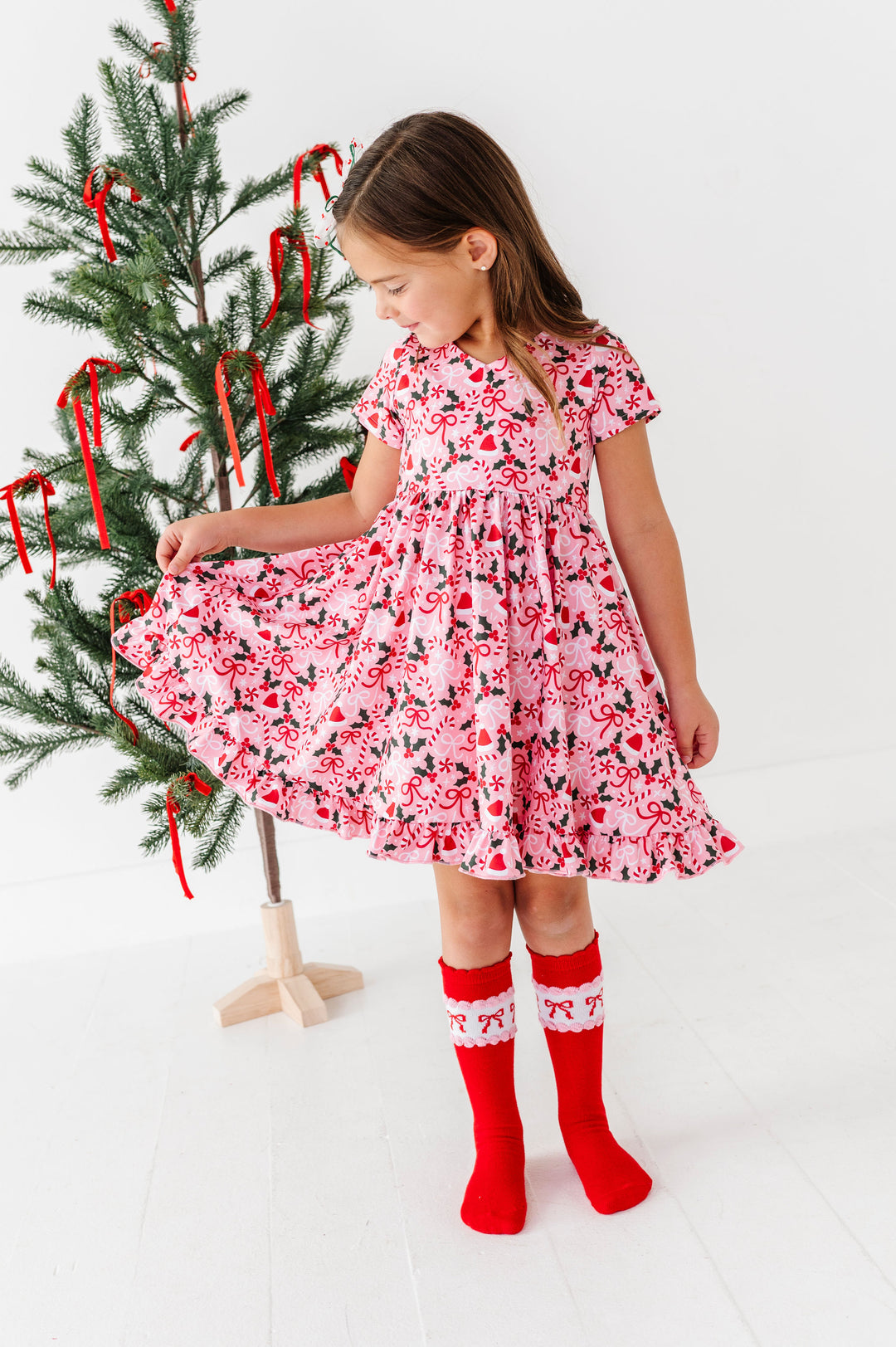 A young girl stands next to a decorated Christmas tree wearing a pink and red floral print dress with red and white knit socks, in a bright white room.