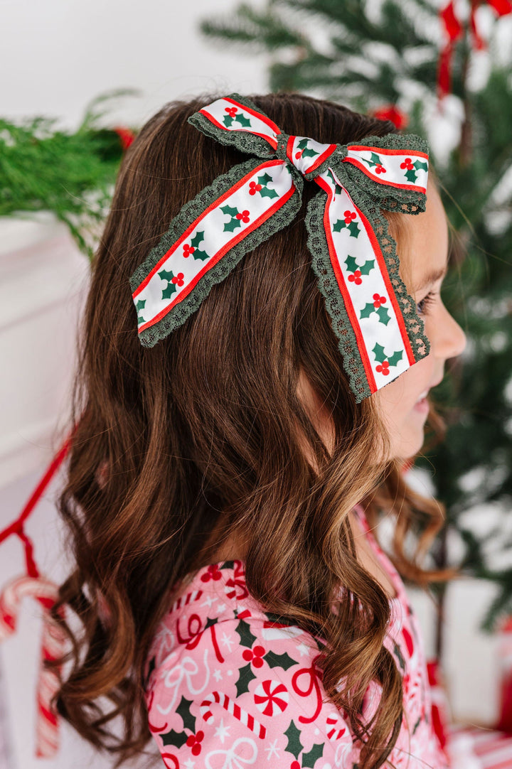 A young girl in profile wearing a festive red, green, and white holiday hair bow headband, standing in front of a decorated Christmas tree and fireplace mantel.