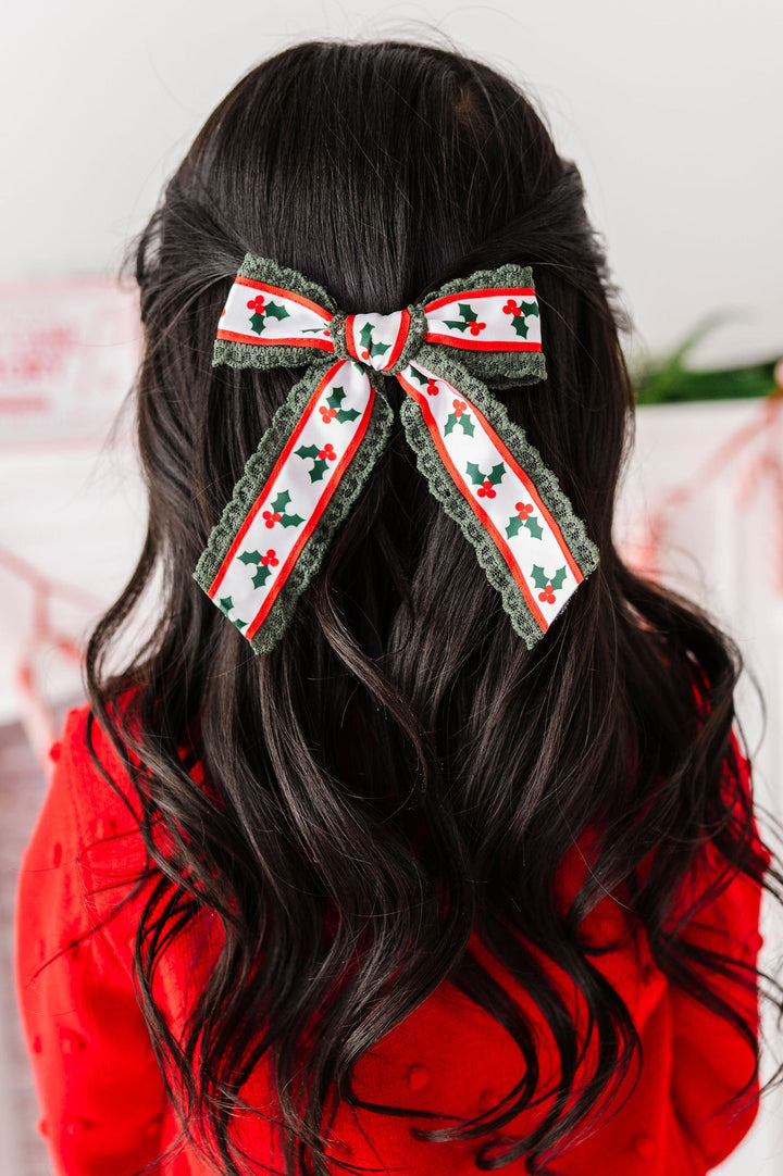 back of little girl with long wavy dark hair and a holly printed ribbon bow in her with dark green lace trim