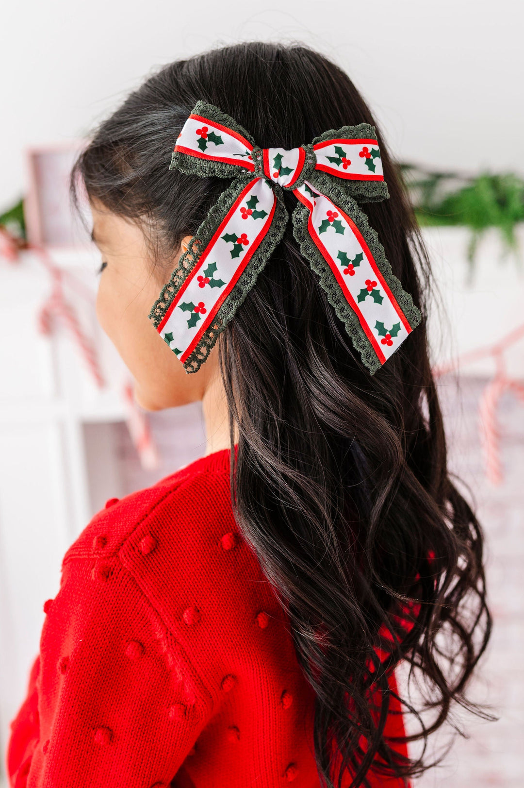 A young girl with long dark hair is shown from the back, wearing a festive red knit sweater and a large holiday-themed hair bow. The bow features a white center with a repeating red and green holly pattern, bordered by olive green lace.