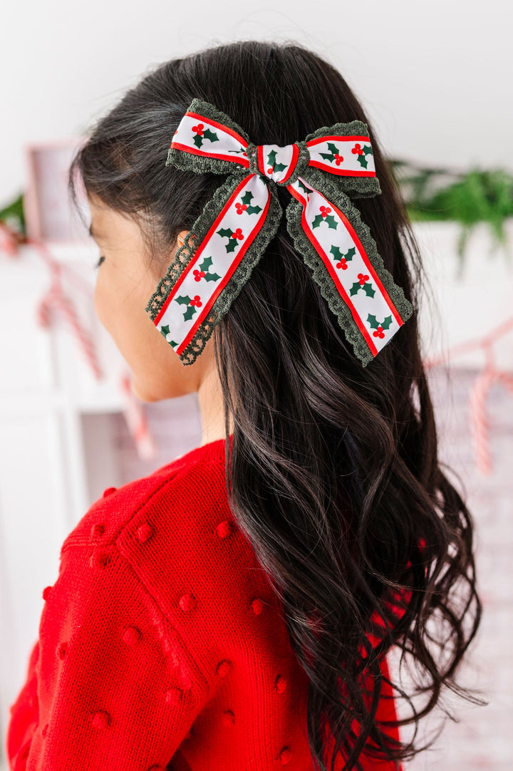 A young girl with long dark hair is shown from the back, wearing a festive red knit sweater and a large holiday-themed hair bow. The bow features a white center with a repeating red and green holly pattern, bordered by olive green lace.