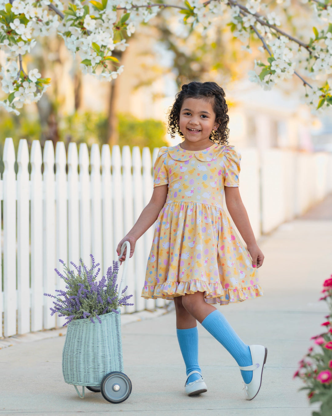 little girl with flower basket wearing pastel yellow collared twirl dress with cute Easter themed print and matching spring blue cable knit knee highs