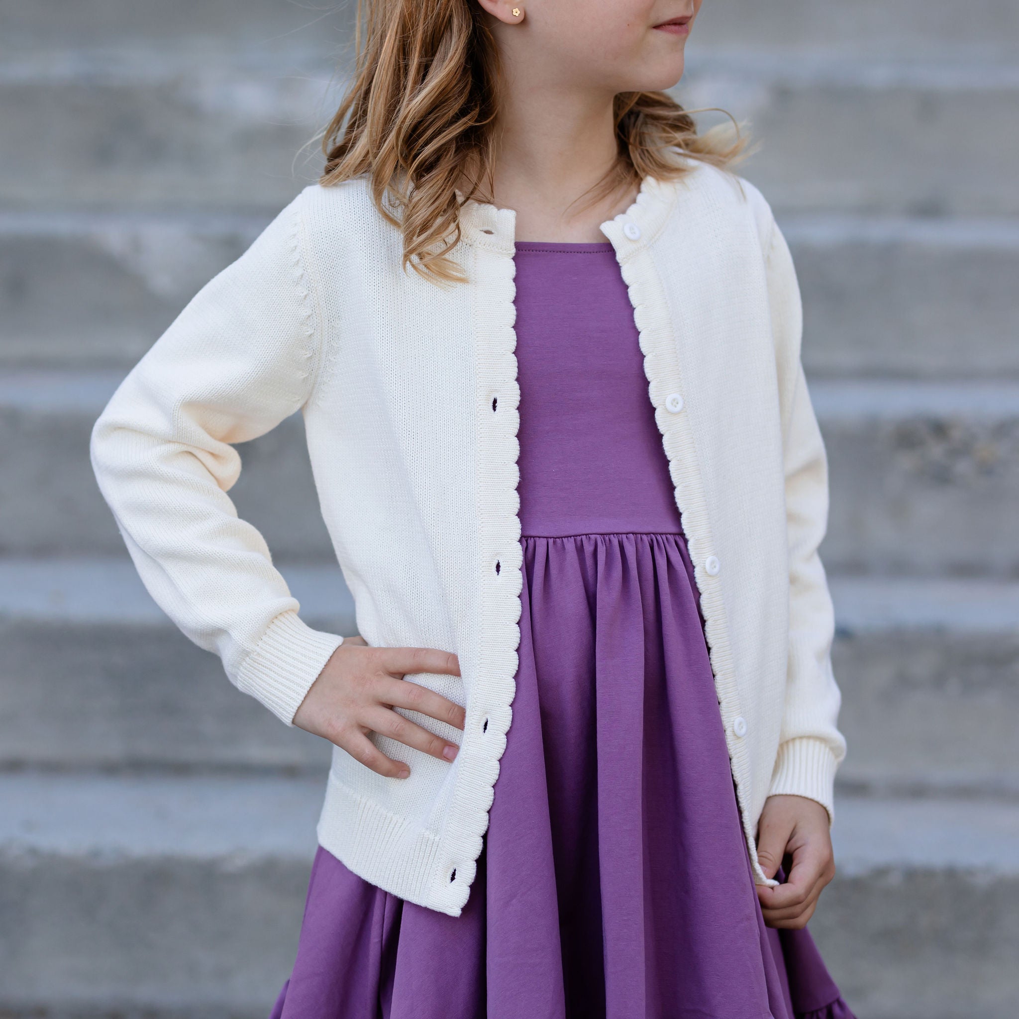 little girl with hand on hip wearing plum purple cotton dress with ivory cardigan sweater over it